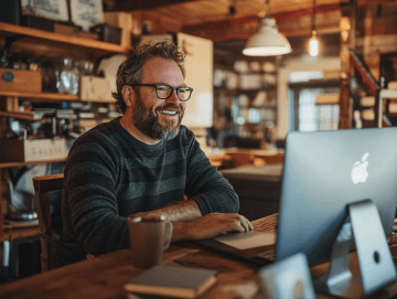 Starší vousatý muž s brýlemi sedí u počítače firmy Apple, sleduje obrazovku a široce se usmívá.